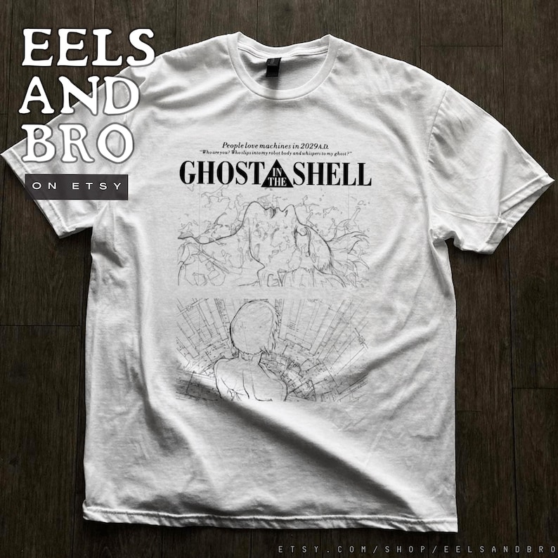 May include: A white short-sleeve t-shirt featuring a black graphic print. The design includes the text GHOST IN THE SHELL, the phrase People love machines in 2029 A.D., and two detailed line art illustrations depicting a figure and a cityscape.