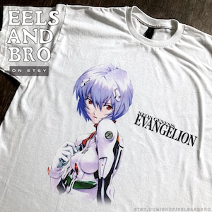 May include: White t-shirt with a vibrant anime character illustration with blue hair and red eyes. The text "NEON GENESIS EVANGELION" is printed on the front. The Etsy shop name "EELS AND BRO" is printed on the shirt.