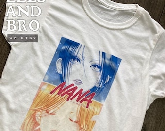 May include: White t-shirt featuring a graphic of a manga character with blue hair and the word "NANA" in red. The image is a close-up of the character's face. The shirt has a crew neck and short sleeves. The Etsy shop name "EELS AND BRO" is printed on the shirt.