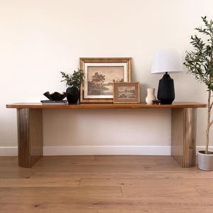 May include: A wooden console table with gold metal legs. The table is decorated with a framed landscape painting, a small framed print, a black vase with greenery, a book, and a white lamp with a black base.