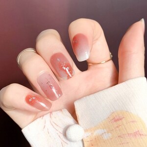 Könnte beinhalten: Eine Hand mit lackierten Nägeln. Die Nägel zeigen einen Farbverlauf von transparenten, rosa und roten Farbtönen. Einige Nägel sind mit silbernem Glitzer und zarten floralen Mustern verziert. Ein goldener Ring ist zu sehen.