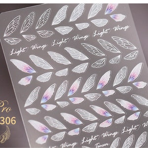 Peut inclure: Feuille transparente d'autocollants pour nail art avec des motifs d'ailes blanches et des reflets irisés. Les mots "Light Wings" sont imprimés en écriture cursive. La feuille porte également la mention "Pro TS-306".