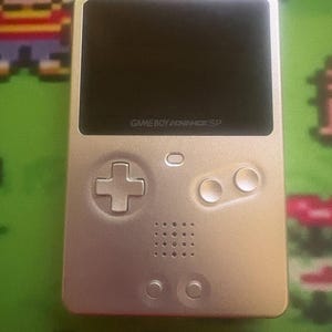 May include: A silver Game Boy Advance SP handheld gaming console featuring a black screen, a directional pad, two circular action buttons, a small status light, a speaker grille, and two smaller circular buttons at the bottom of the device.
