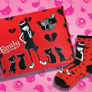 May include: A red box with the words "Emily the Strange" on the front. The box has a broken heart design and a black and white illustration of Emily the Strange. The box is sitting on a pink background with black cat illustrations. A pair of red socks with a black and white illustration of Emily the Strange on them are in the foreground.