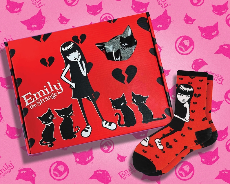 May include: A red box with the words "Emily the Strange" on the front. The box has a broken heart design and a black and white illustration of Emily the Strange. The box is sitting on a pink background with black cat illustrations. A pair of red socks with a black and white illustration of Emily the Strange on them are in the foreground.