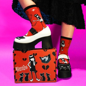 May include: A pair of black and white platform shoes with a black cat graphic on the toe. The shoes are worn with red and black socks featuring a cartoon character playing a guitar. The socks are from the Emily the Strange brand. The shoes are sitting on a red box with a broken heart pattern and a cartoon character on the front. The box says "Emily the Strange" on the front.