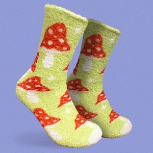 以下が含まれることがあります： 明るい緑色のふわふわソックスのペア。赤と白のキノコの繰り返し模様が施されています。ソックスは白い袖口で、明るい青色の背景に配置されています。