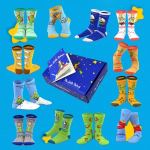 K&ouml;nnte beinhalten: Ein Set mit 10 Paar bunten Socken mit Illustrationen des kleinen Prinzen. Die Socken sind in einer blauen Box mit einem Buch verpackt, auf der der Titel "Der kleine Prinz" steht.