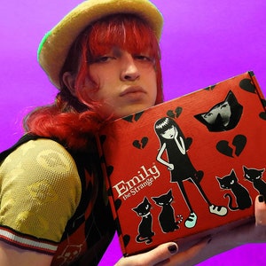 May include: A red box with the words "Emily the Strange" on the front. The box features a black and white illustration of a girl with black hair and a black dress, along with several black cats. The box is held by a person wearing a yellow and black vest.