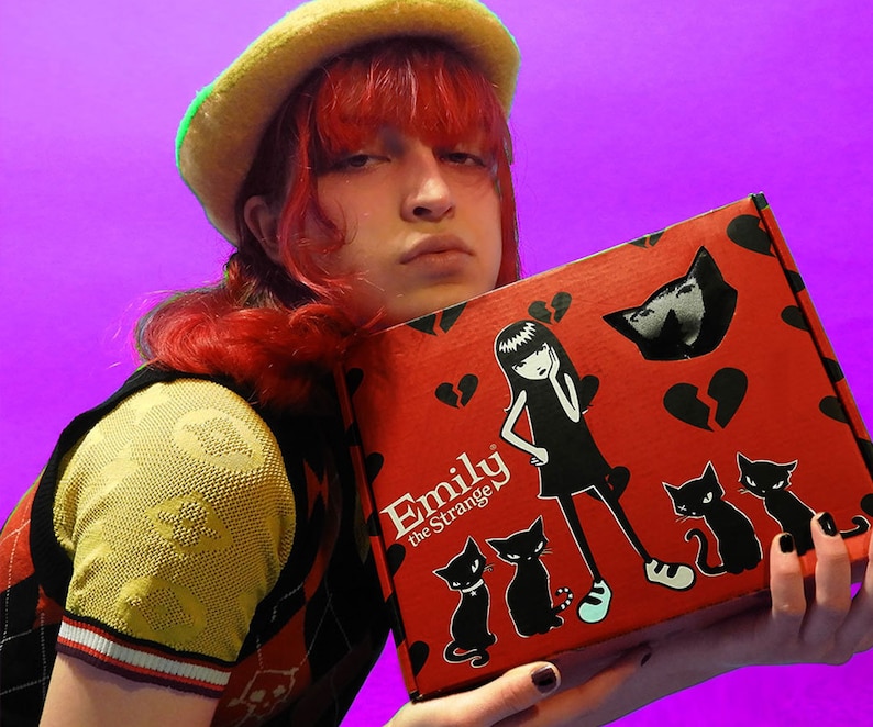 May include: A red box with the words "Emily the Strange" on the front. The box features a black and white illustration of a girl with black hair and a black dress, along with several black cats. The box is held by a person wearing a yellow and black vest.