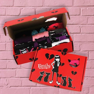 Puede incluir: Una caja roja con un diseño de corazón roto y el texto "Emily the Strange" en la parte delantera. La caja contiene un juego de calcetines con gatos negros y otros diseños.