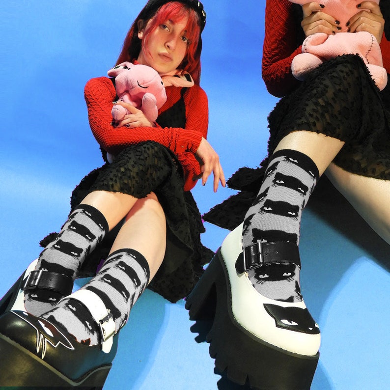 May include: A person wearing black and white platform shoes with a black cat print on the socks and shoes. The person is wearing a red sweater and a black skirt. The person is holding a pink stuffed animal.