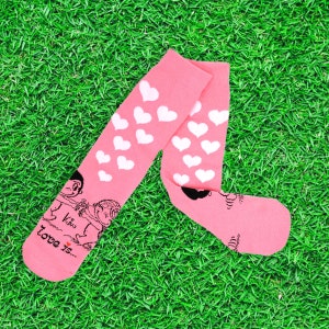 Puede incluir: Calcetines rosas con patrones de corazones blancos y una ilustración de dibujos animados en blanco y negro. El texto "Kiss Love is..." también es visible. Los calcetines están sobre una superficie verde.