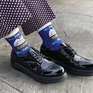 K&ouml;nnte beinhalten: Ein Paar schwarze Lackleder-Brogue-Schuhe mit wei&szlig;en Schn&uuml;rsenkeln. Die Schuhe werden mit blauen Socken getragen, die ein wei&szlig;es Motiv eines Polizeihutes und den Text "TR.a fun... UN.a.doubt... TR.a fun" zeigen.