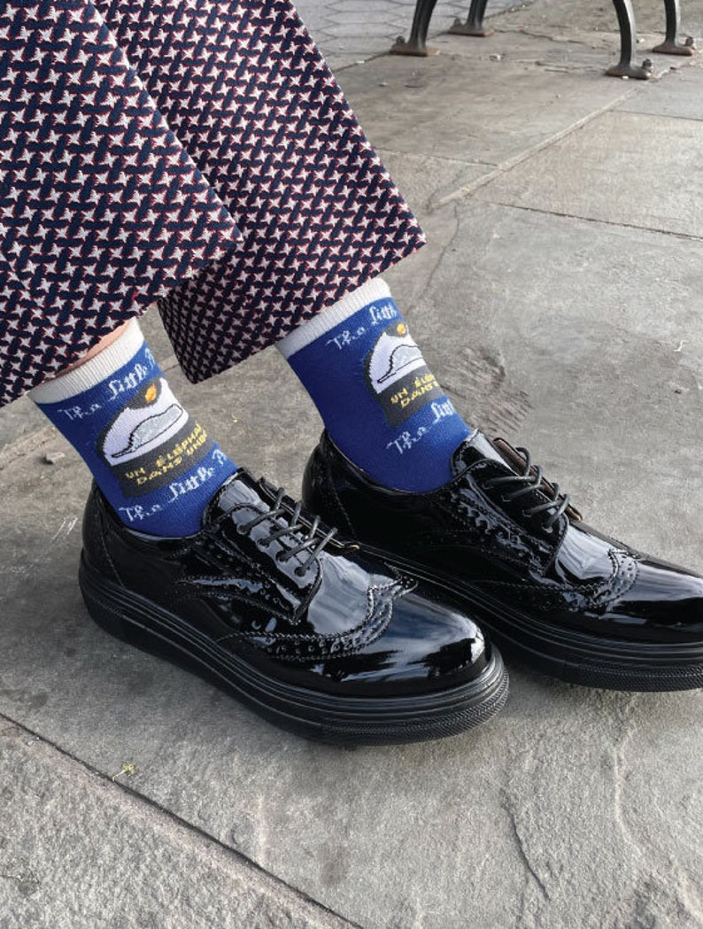 K&ouml;nnte beinhalten: Ein Paar schwarze Lackleder-Brogue-Schuhe mit wei&szlig;en Schn&uuml;rsenkeln. Die Schuhe werden mit blauen Socken getragen, die ein wei&szlig;es Motiv eines Polizeihutes und den Text "TR.a fun... UN.a.doubt... TR.a fun" zeigen.