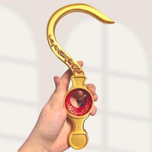 Puede incluir: Un accesorio en forma de gancho de color dorado con una sección superior curva y una base circular con un orificio central. La superficie está decorada con intrincados patrones de filigrana roja a lo largo del brazo curvo y el centro.