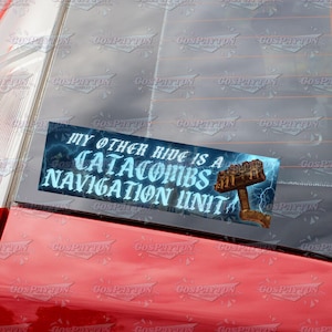 Puede incluir: Una pegatina rectangular con el texto "MY OTHER RIDE IS A CATACOMBS NAVIGATION UNIT" en azul claro, con un gráfico de un martillo. La pegatina está pegada a un coche rojo.