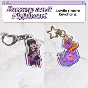 Könnte beinhalten: Zwei Schlüsselanhänger aus Acryl mit den Motiven Buzzy und Figment. Der Buzzy-Schlüsselanhänger ist eine Comicfigur mit einem grünen Hut und einem blauen Hemd. Der Figment-Schlüsselanhänger ist ein lila Drache mit einer gelben Mähne. Beide Schlüsselanhänger haben einen silbernen Ring und einen goldenen Sternanhänger.