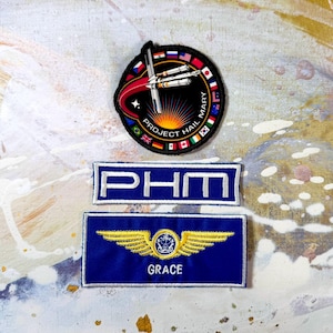 May include: A circular embroidered patch featuring a spacecraft, the text PROJECT HAIL MARY, and international flags, alongside a rectangular blue patch with the text PHM and a rectangular blue patch with gold wings and the name GRACE.