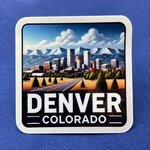 Op de afbeelding: Een vierkante sticker met een schilderachtig uitzicht op Denver, Colorado. De afbeelding toont een stadsgezicht met wolkenkrabbers, een weg en bergen op de achtergrond. De woorden "DENVER" en "COLORADO" zijn wit gedrukt.