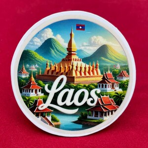 Op de afbeelding: Ronde witte sticker met een gedetailleerde illustratie van een gouden tempel en traditionele gebouwen tegen een achtergrond van groene bergen en een blauwe lucht. Het woord "Laos" is in witte letters geschreven.