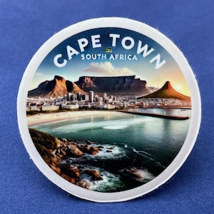 Op de afbeelding: Ronde sticker met een landschap van Kaapstad, Zuid-Afrika. De afbeelding toont een kustlijn met een stad, bergen en de zee. De tekst "CAPE TOWN SOUTH AFRICA" is in een boog bovenaan gedrukt.
