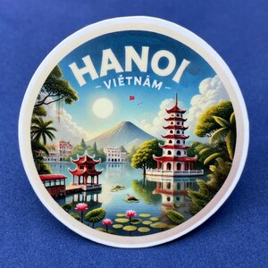 May include: Circular sticker with a scenic illustration of Hanoi, Vietnam. The design features a pagoda, lake, mountain, and the text "HANOI - VIETNAM" in white. The sticker has a white border and a blue background.