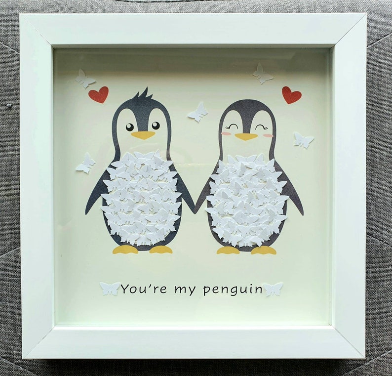 May include: A framed artwork featuring two cartoon penguins holding hands, with white butterfly accents and red heart details. The text "You're my penguin" is at the bottom. The frame is white.