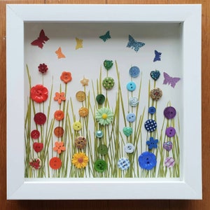 May include: A framed artwork featuring a rainbow of colourful buttons arranged in a field of green grass. The buttons are shaped like flowers, stars, and hearts. There are also butterflies and other decorative elements.