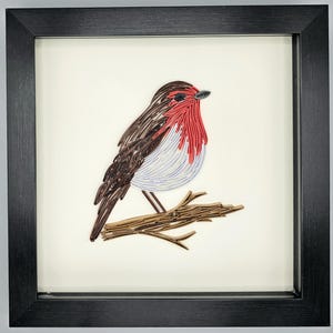 May include: Framed artwork featuring a detailed paper quilling of a robin perched on a branch. The bird has a red breast, brown and white wings, and is set against a cream background. The frame is black.
