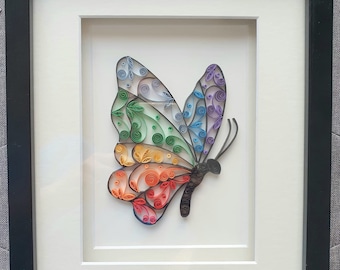 May include: A framed paper craft of a colorful butterfly with rainbow wings. The butterfly is made of curled paper and has a detailed design.