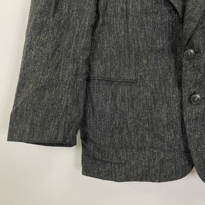 Op de afbeelding: Een donkergrijze blazer van een getextureerde stof. De blazer heeft een enkele knoopsluiting en een zak aan de linkerkant.