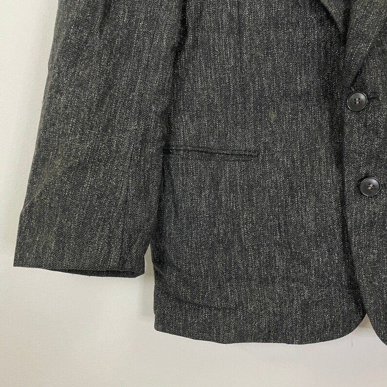 Op de afbeelding: Een donkergrijze blazer van een getextureerde stof. De blazer heeft een enkele knoopsluiting en een zak aan de linkerkant.