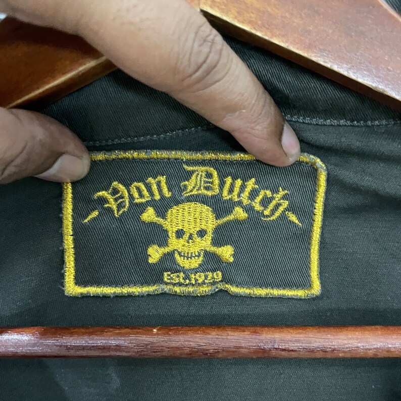 May include: A close-up of a dark green fabric patch with yellow stitching. The patch features a skull and crossbones with the text "Von Dutch" and "Est. 1929".