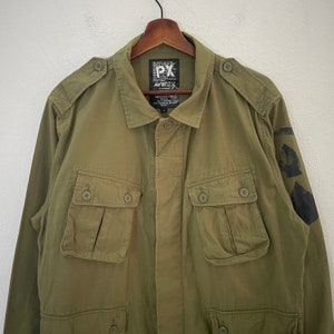 Op de afbeelding: Olijfgroene jas met meerdere zakken met knopen en schouderstukken. De jas heeft een knoopsluiting aan de voorkant en een kraag. De merknaam "AVIREX" is zichtbaar op een label bij de kraag. Zwarte patches zitten op de linkermouw.