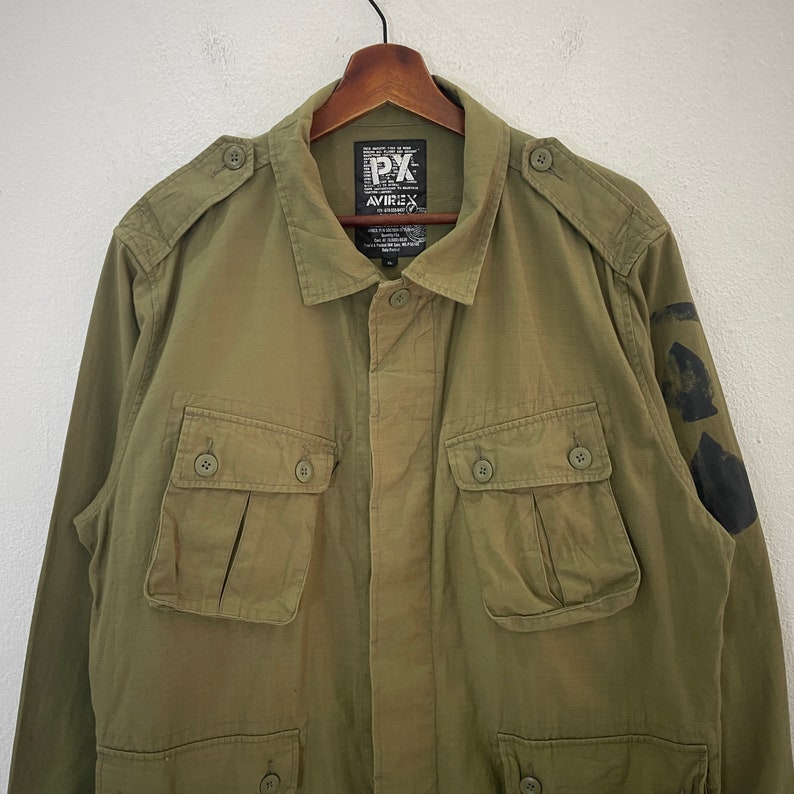 Op de afbeelding: Olijfgroene jas met meerdere zakken met knopen en schouderstukken. De jas heeft een knoopsluiting aan de voorkant en een kraag. De merknaam "AVIREX" is zichtbaar op een label bij de kraag. Zwarte patches zitten op de linkermouw.
