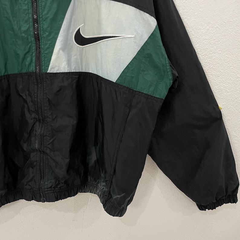 May include: Black, green, and silver Nike windbreaker jacket. The jacket features a full zipper, a white Nike swoosh logo, and color-blocked panels. The jacket is made of a lightweight, water-resistant material.