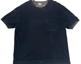 May include: Dark navy blue short-sleeved t-shirt with a chest pocket. The shirt has a crew neck and rolled-up sleeves. The brand logo is visible on the shirt and the collar. The shirt is hanging on a wooden hanger.