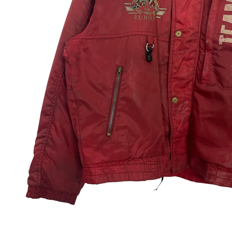 K&ouml;nnte beinhalten: Rote Vintage-Jacke mit Rei&szlig;verschlusstasche und kleinem Emblem &uuml;ber der Tasche. Die Jacke hat einen Knopfverschluss und einen Kordelzug am Saum. Das Wort "EUROPE" ist auf dem Emblem zu sehen.