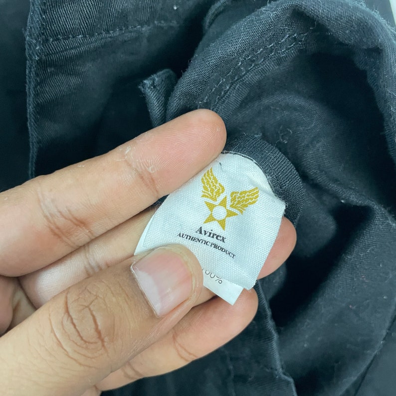 May include: Close-up of a black garment with a white tag. The tag features the brand name "Avirex" in black text, along with a gold emblem of wings and a star. The words "AUTHENTIC PRODUCT" are also printed on the tag.