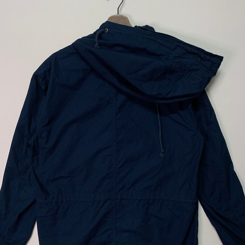 May include: A navy blue hooded jacket with a drawstring hood and waist. The jacket is hanging on a wooden hanger against a white background. The jacket is made of a lightweight fabric.