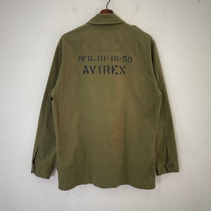 Op de afbeelding: Olijfgroene jas met lange mouwen en een knoopsluiting. De achterkant van de jas heeft de tekst "MFG. 10-16-59" boven het woord "AVIREX" in zwart. De jas hangt aan een houten hanger.