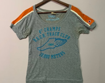 May include: Gray Champion t-shirt with orange and white stripes on the sleeves. The shirt features a blue graphic with the text "6th Champs U.S.C.H. Track Club 1964 10,000 Meters."