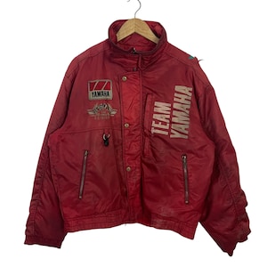 K&ouml;nnte beinhalten: Eine rote Vintage Yamaha Team Jacke mit hohem Kragen und mehreren Taschen. Die Jacke hat das Yamaha-Logo und die Worte "TEAM YAMAHA" in Wei&szlig; aufgedruckt. Sie hat einen Rei&szlig;verschluss und Druckkn&ouml;pfe.