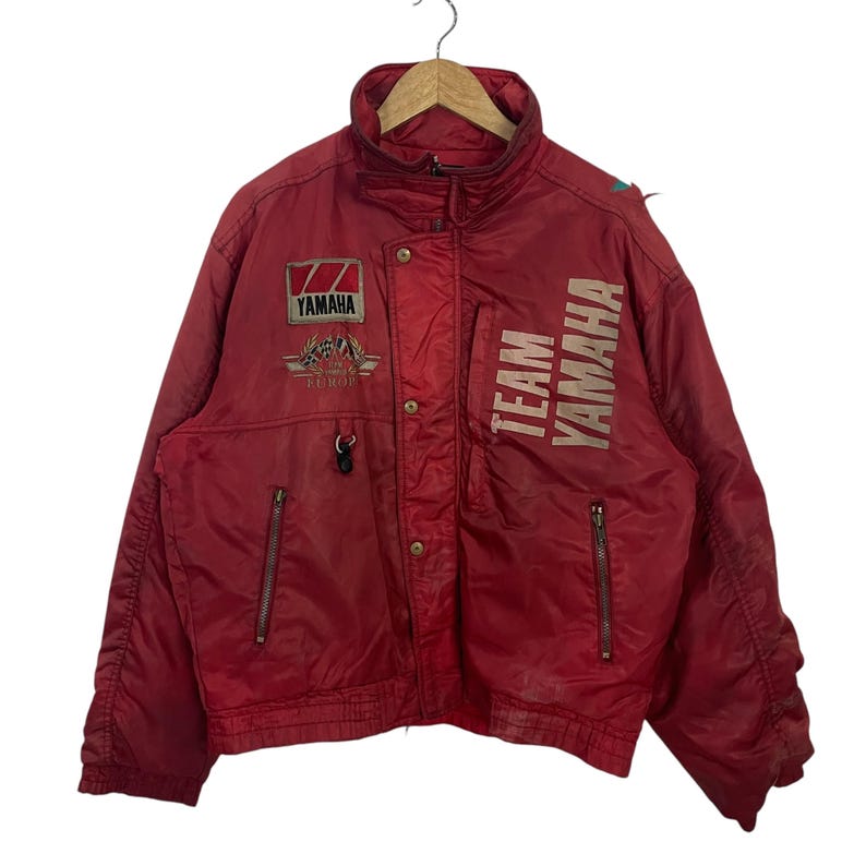 K&ouml;nnte beinhalten: Eine rote Vintage Yamaha Team Jacke mit hohem Kragen und mehreren Taschen. Die Jacke hat das Yamaha-Logo und die Worte "TEAM YAMAHA" in Wei&szlig; aufgedruckt. Sie hat einen Rei&szlig;verschluss und Druckkn&ouml;pfe.