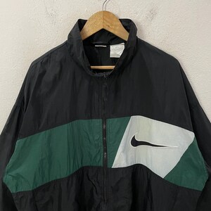 May include: Black and green Nike windbreaker jacket with a white Nike swoosh logo. The jacket has a full zipper and a high collar. The design includes a green horizontal stripe across the chest and a white and gray panel with the Nike logo.