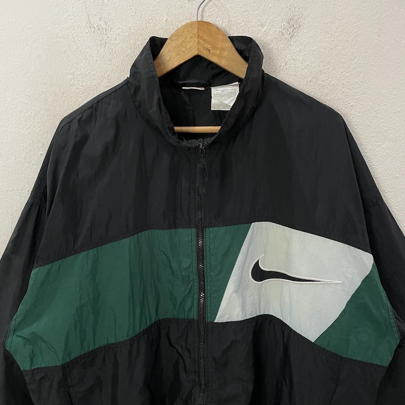 May include: Black and green Nike windbreaker jacket with a white Nike swoosh logo. The jacket has a full zipper and a high collar. The design includes a green horizontal stripe across the chest and a white and gray panel with the Nike logo.