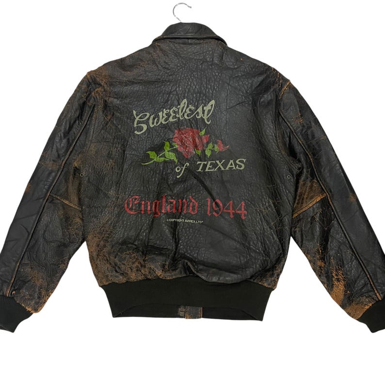 May include: A dark brown distressed leather bomber jacket with a black ribbed hem and cuffs. The back features a red rose graphic with the text Sweetest of TEXAS and England 1944 printed in red and white lettering, including a small copyright notice.