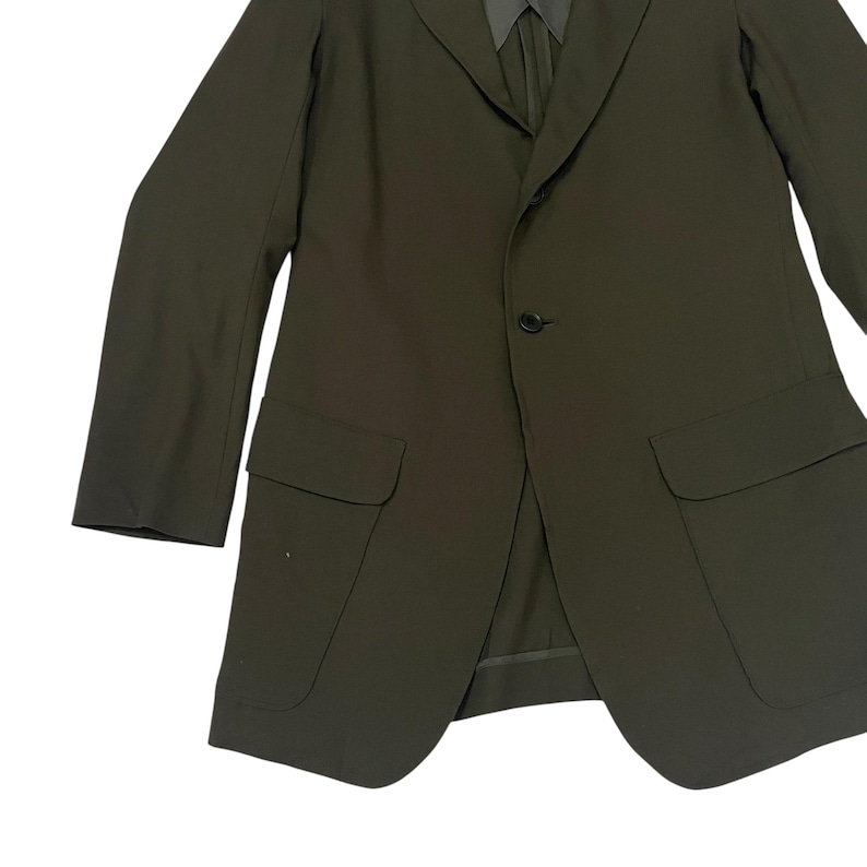 Puede incluir: Una americana de color verde oliva oscuro con solapa de muesca, cierre de un solo bot&oacute;n en la parte delantera y dos grandes bolsillos de parche con solapa en la parte inferior. La chaqueta tiene mangas largas y un cuello estructurado mate.