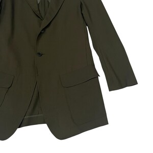 Puede incluir: Un blazer de color verde oliva con solapa de muesca, dos botones oscuros para el cierre y dos grandes bolsillos con solapa en la parte delantera. La prenda tiene mangas largas y un dise&ntilde;o estructurado con un acabado de tela lisa.